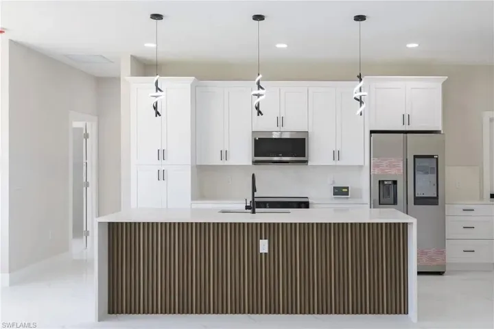 Kitchen with stainless steel appliances, an island with sink, white cabinetry, light stone counters, and decorative light fixtures