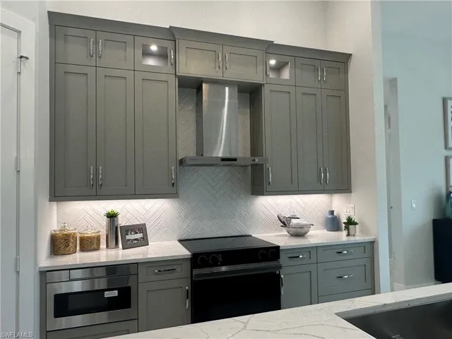 Kitchen with light stone counters, wall chimney range hood, built in microwave, tasteful backsplash, and electric range