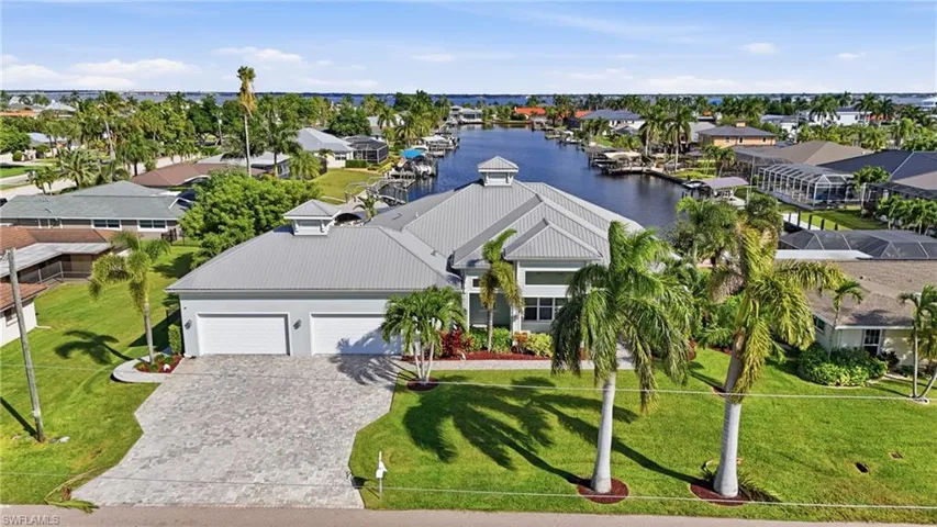 Waterfront residence featuring a metal roof, cupola accents, paver driveway, three-car garage, and established tropical landscaping