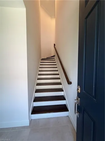 Entryway featuring a dark paneled door with brushed nickel hardware, light-toned tile flooring, and stairs with dark wood-finish treads and white risers, complemented by a wood-finish handrail and crown molding