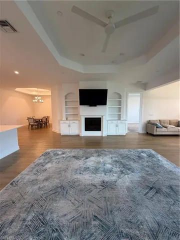 Living area featuring wood-finish flooring, a tray ceiling with a ceiling fan, recessed lighting, and a fireplace with built-in shelving and cabinetry