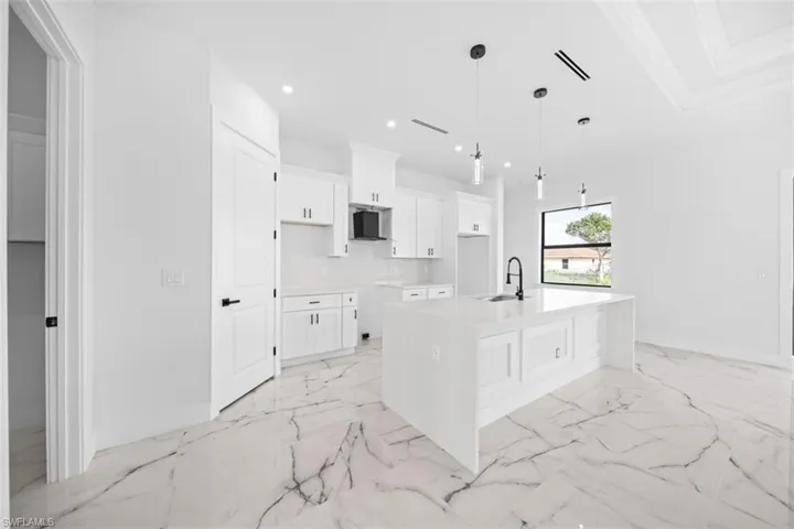 Open-concept kitchen featuring white cabinetry, a large center island with a built-in sink, pendant lighting, and white flooring with dark veining