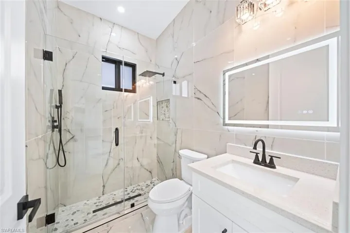 Spacious bathroom featuring large format stone-look tile walls, a glass-enclosed shower with pebble-tile flooring, a vanity with a light-toned countertop, and an illuminated mirror