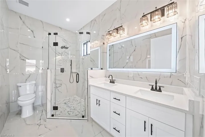 Dual vanity with white cabinetry and integrated sinks, illuminated mirror, walk-in shower with clear glass enclosure, marble-patterned tile, and recessed lighting