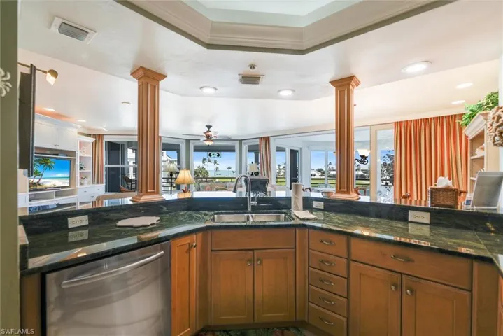 Kitchen with decorative columns, stainless steel dishwasher, wood finish cabinets, dark stone countertops, and healthy amount of natural light