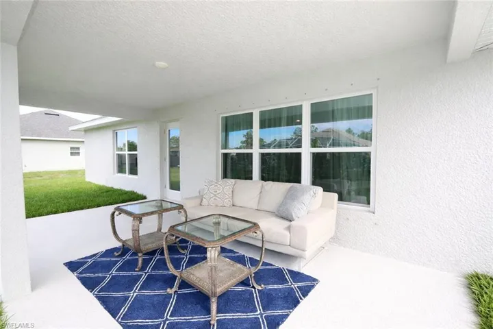 Covered outdoor patio area featuring a white textured wall, large windows, and a view of the surrounding green space