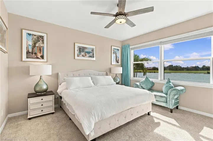 Neutral-toned bedroom featuring plush wall-to-wall carpeting, a multi-blade ceiling fan with integrated lighting, and a large horizontal window with water and landscape scenery