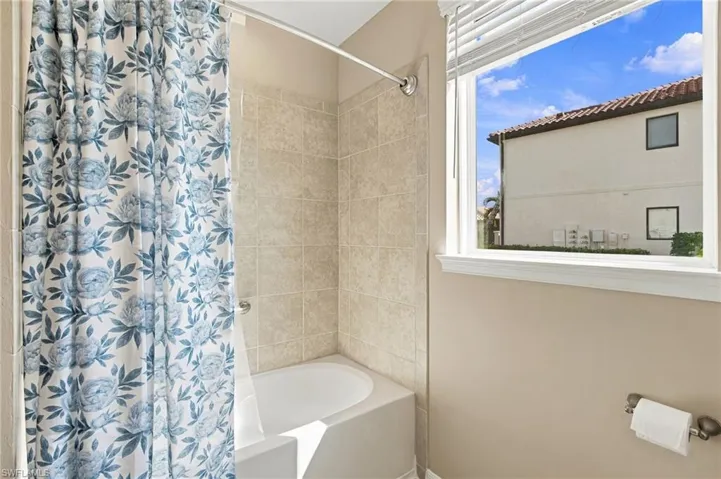 Bathtub enclosure featuring neutral ceramic wall tile, polished metal shower rod, and a window with integrated horizontal blinds