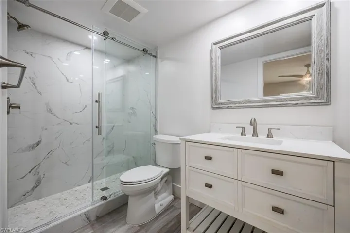 Bathroom featuring a walk-in shower with clear glass enclosure and marble-look tile, a white vanity with integrated sink and brushed nickel hardware, and wood-finish flooring