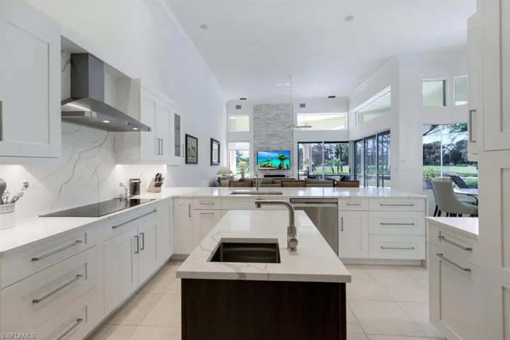 Kitchen with black electric stovetop, wall chimney range hood, an island with sink, stainless steel dishwasher, and light stone counters