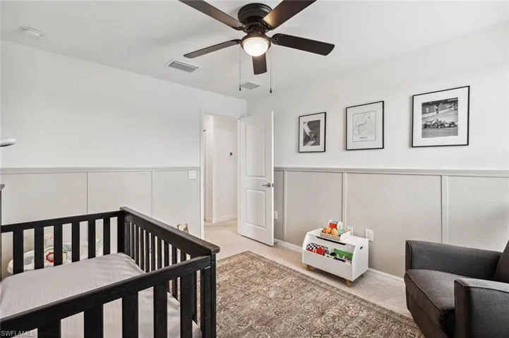 Bedroom featuring a decorative wall, a nursery area, wainscoting, ceiling fan, and carpet flooring