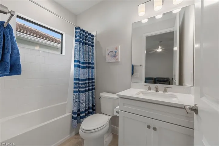 Bathroom featuring a white vanity cabinet with a light-toned solid surface countertop, rectangular under-mount sink, polished chrome faucet, full-width mirror with integrated lighting, and a white tile shower surround