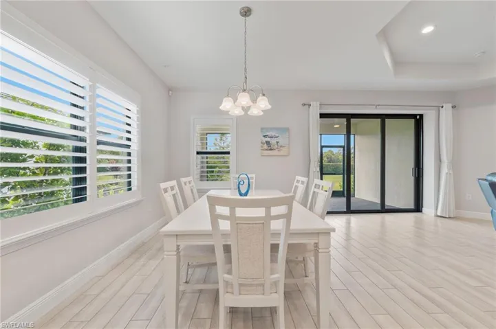 Light-filled interior featuring wood-finish tile flooring, recessed tray ceiling, multi-pane chandelier, plantation shutters, and black-framed sliding glass doors