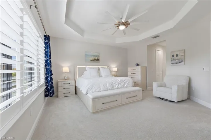 Tray ceiling with a multi-blade ceiling fan, expansive windows featuring plantation shutters, and neutral wall-to-wall carpeting