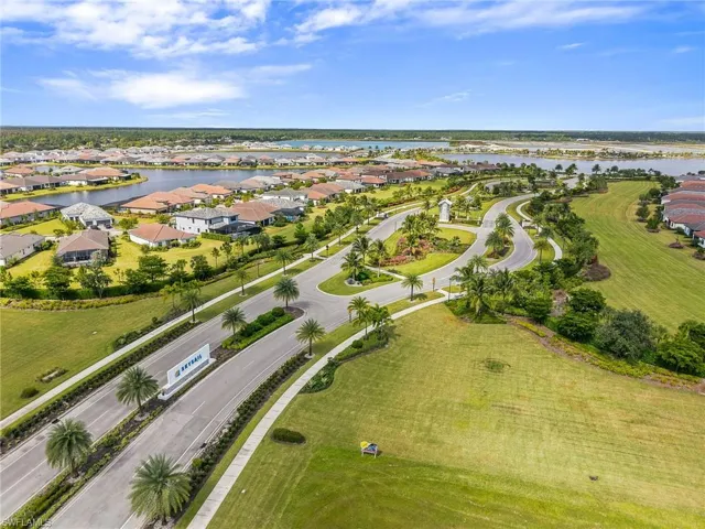 Aerial perspective of a community featuring asphalt roadways, landscaped center medians, mature palm trees, sprawling green lawns, and water frontage
