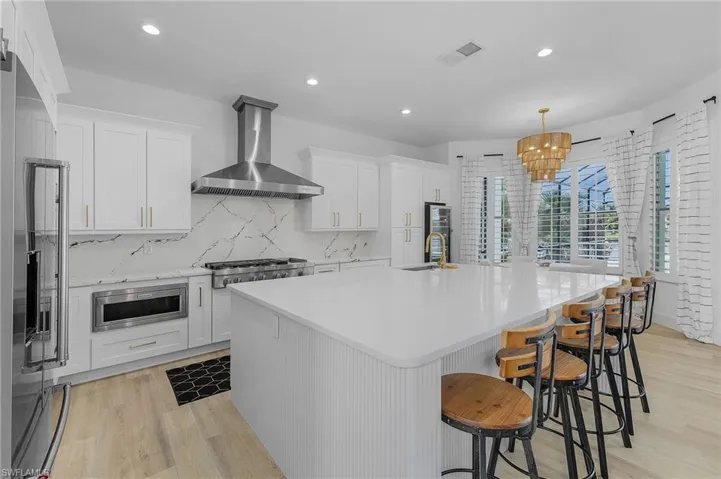 Kitchen with a kitchen island with sink, range hood, appliances with stainless steel finishes, decorative light fixtures, and white shaker cabinetry