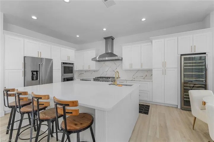 Kitchen with white shaker cabinets, wine cooler, exhaust hood, an island with sink, and stainless steel appliances