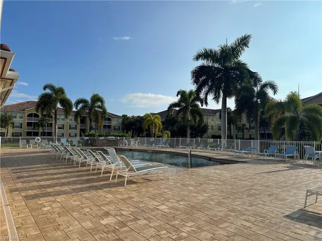 Expansive paver patio surrounding an in-ground swimming pool, bordered by a white metal fence and lush palm trees