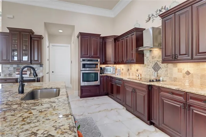 Kitchen with wall chimney range hood, stainless steel double oven, black electric cooktop, reddish brown cabinets, and ornamental molding