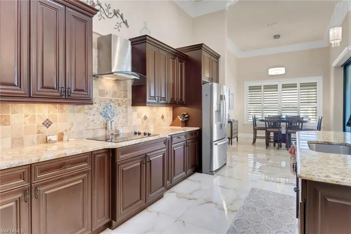 Kitchen with stainless steel fridge, wall chimney exhaust hood, black electric stovetop, light marble finish floors, and decorative backsplash