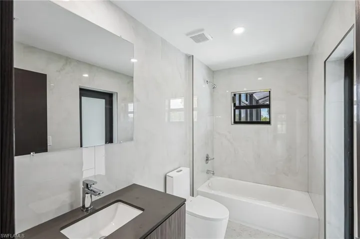 Modern bathroom featuring a rectangular vanity with a dark countertop and integrated sink, a large wall-mounted mirror, a tub/shower combination with chrome fixtures, and light-toned wall tiling
