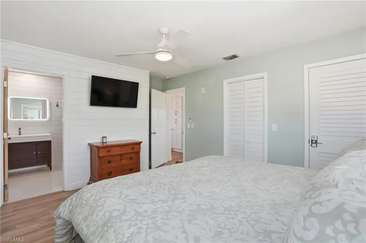 Bedroom featuring light wood-style floors, two closets, ceiling fan, wooden walls, and ensuite bath - Virtually Edited Image