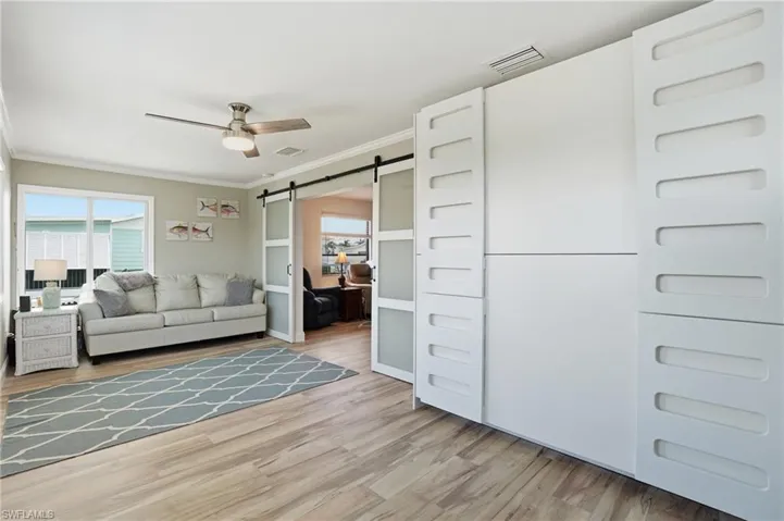 Den featuring a barn door, crown molding, ceiling fan, plenty of natural light, and light wood-style floors - Virtually Edited Image