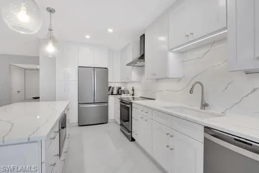 Kitchen featuring wall chimney exhaust hood, light stone counters, hanging light fixtures, stainless steel appliances, and white cabinetry