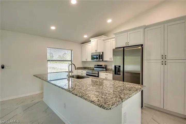 Modern kitchen featuring white cabinetry, stainless steel appliances including a double-door refrigerator and built-in microwave, granite-style countertops, a spacious center island with seating, recessed ceiling lighting, and tile flooring throughout the space