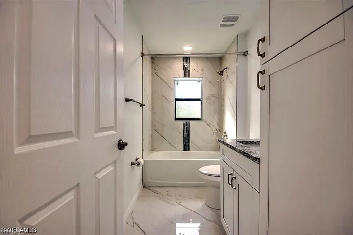 White paneled interior door, white vanity with stone countertop, white tiled floor, white bathtub with tiled surround, and a fixed window