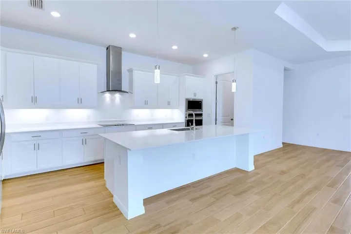 Kitchen featuring wall chimney range hood, pendant lighting, white cabinetry, a kitchen island with sink, and light wood finished floors