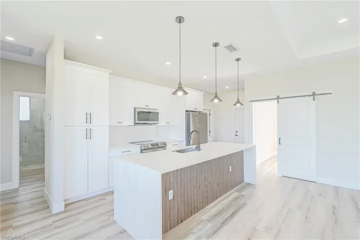 Kitchen featuring a barn door, decorative light fixtures, an island with sink, white cabinetry, and stainless steel appliances