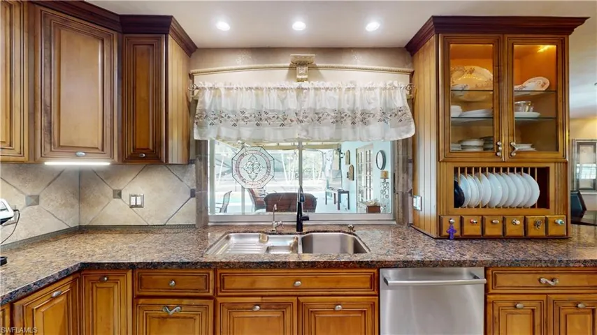 Kitchen with brown cabinetry, stainless steel dishwasher, dark stone counters, backsplash, and glass insert cabinets