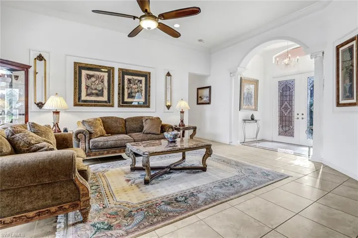 Living room with crown molding, ceiling fan with notable chandelier, light tile floors, and decorative columns