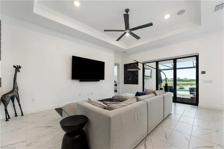 Spacious living area featuring large format white tile flooring with grey veining, a tray ceiling with crown molding, and a matte black ceiling fan