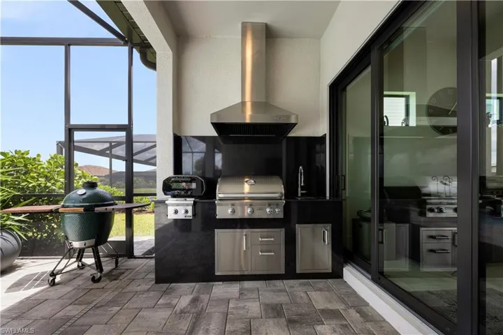 Outdoor kitchen featuring a built-in stainless steel grill, integrated sink, stainless steel under-counter cabinetry, a stainless steel range hood, and a dark countertop with a matching backsplash
