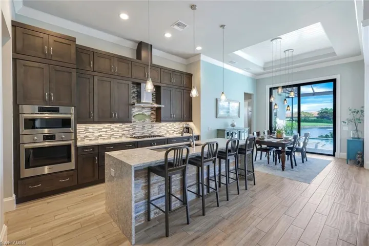 Kitchen featuring a raised ceiling, hanging light fixtures, a kitchen island with sink, wall chimney range hood, and a breakfast bar