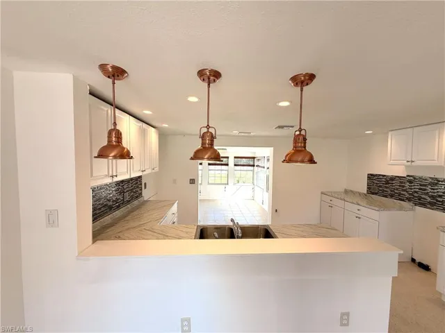 Kitchen featuring three copper-finish pendant lights, stone-patterned countertops, white shaker-style cabinetry, a dual-basin stainless steel sink, and a multi-toned horizontal tile backsplash