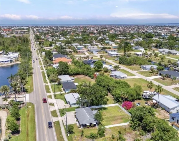 Aerial perspective of a residential lot featuring a paved driveway, established landscaping, white perimeter fencing, and proximity to a canal