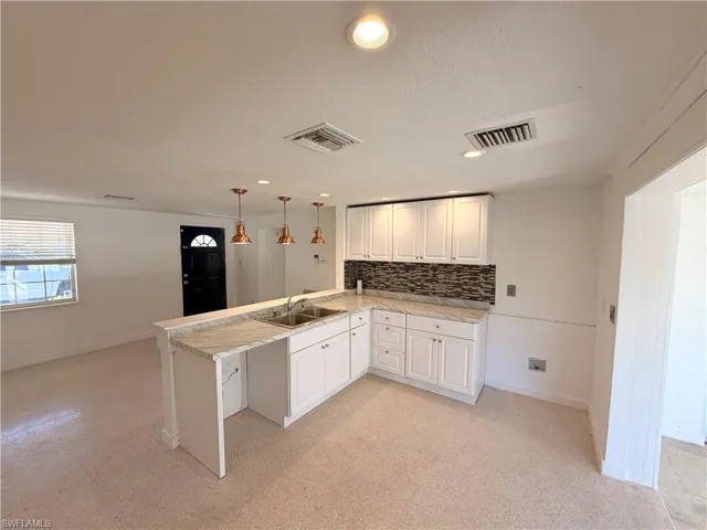 Kitchen featuring white cabinetry, stone-patterned countertops, a stainless steel double-basin sink, multi-tone tile backsplash, and decorative pendant lighting