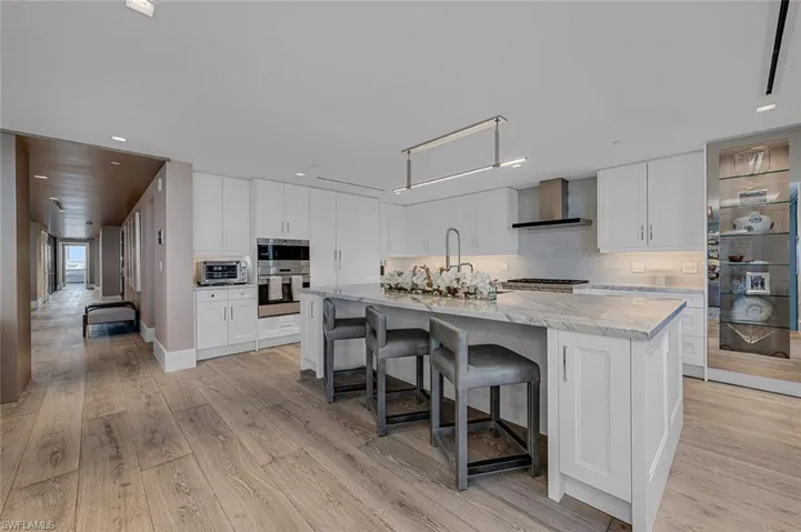Kitchen with wall chimney range hood, an island with sink, light wood-type flooring, and white cabinets