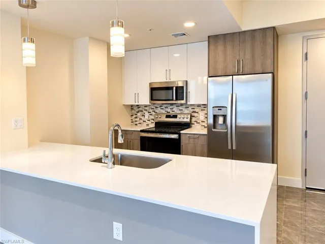 Kitchen featuring an island with a quartz countertop and integrated sink, stainless steel appliances, multi-toned cabinetry, a mosaic tile backsplash, and overhead pendant lighting