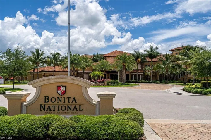 Gated community entrance with an architectural sign, lush landscaping, and paver driveways leading to a large building with a terracotta tile roof and extensive palm trees