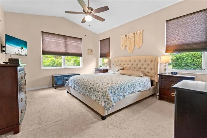 Spacious primary bedroom featuring vaulted ceilings, neutral-toned carpeting, multiple windows with privacy blinds, and a ceiling fan with integrated lighting