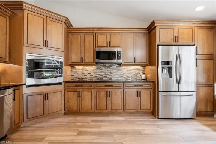 Kitchen with tasteful backsplash, under cabinet lighting, stainless steel appliances, dark stone countertops, vaulted ceiling, and light plank tile flooring