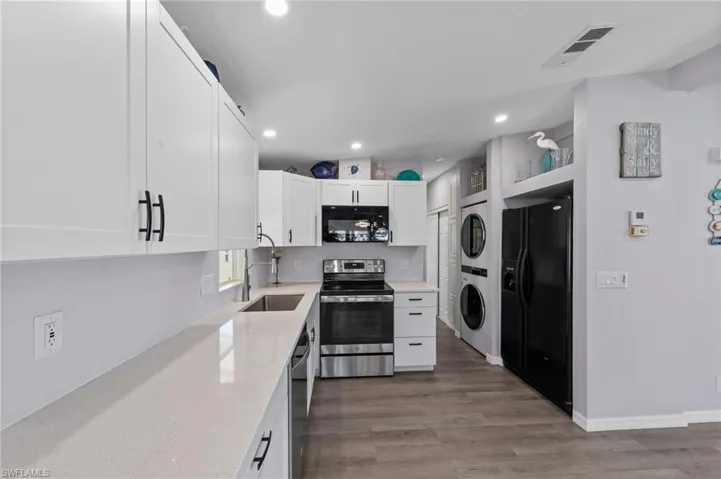 Kitchen with black appliances, stacked washer / drying machine, white cabinets, light stone counters, and light wood-style flooring