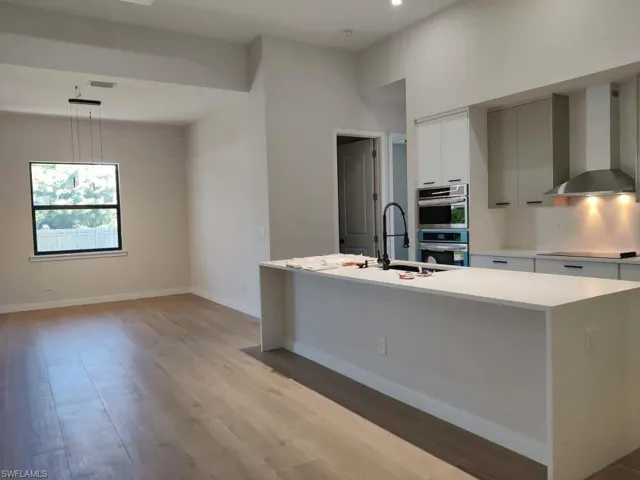 Kitchen featuring wall chimney range hood, light wood-type flooring, double oven, hanging light fixtures, and light stone counters