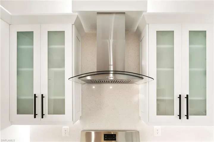 Kitchen view of wall chimney range hood, glass insert cabinets, white cabinets, and light stone countertops