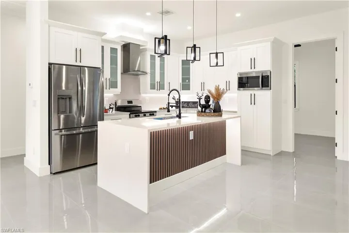 Kitchen with glass insert cabinets, appliances with stainless steel finishes, wall chimney exhaust hood, light stone countertops, and an island with sink
