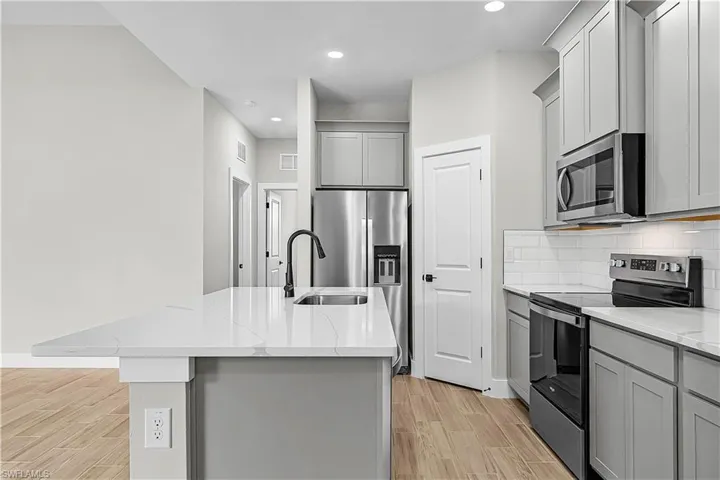 Kitchen with stainless steel appliances, gray cabinets, wood finish floors, a center island with sink, and light stone countertops